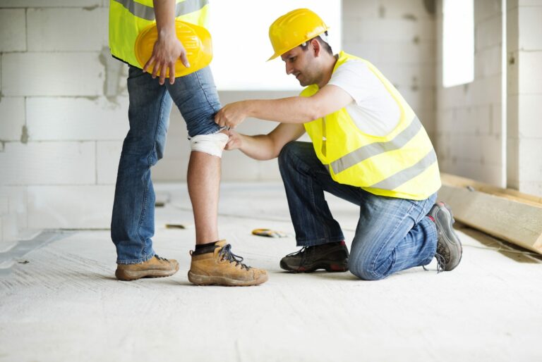 Ein am Knie verletzter Bauarbeiter, der von einem Kollegen mit einem Verband versorgt wird.