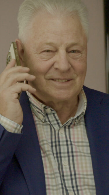 Ein älterer Mann lächelt in die Kamera während er mit einem Smartphone am Ohr telefoniert.