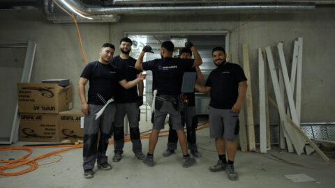 Eine Gruppe von Azubis der Firma elektroNWS ist abgebildet. Sie stehen in einer Tiefgarage beieinander und lächeln, während sie auf einen Azubi in der Mitte zeigen.